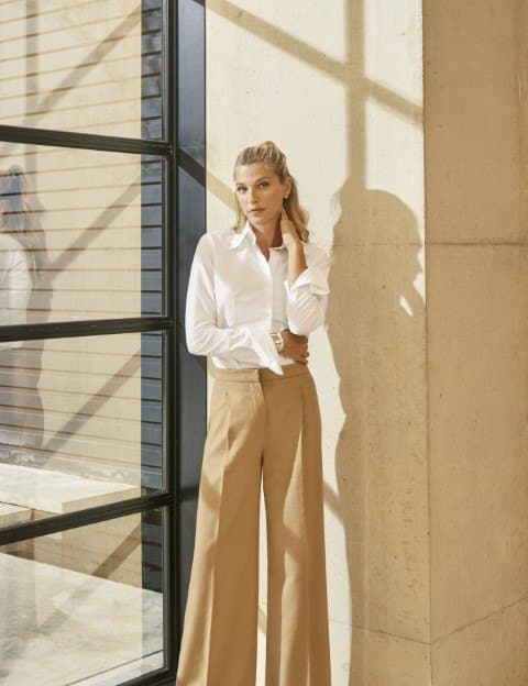 Nicole Junkermann in a white shirt beside a tall window, standing in bright daylight against a pale stone wall